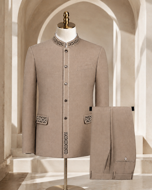 A beige formal jacket displayed on a mannequin with matching trousers folded beside it, set against an arched interior backdrop. The jacket has decorative embroidered detailing at the collar, front placket, and pocket flaps.